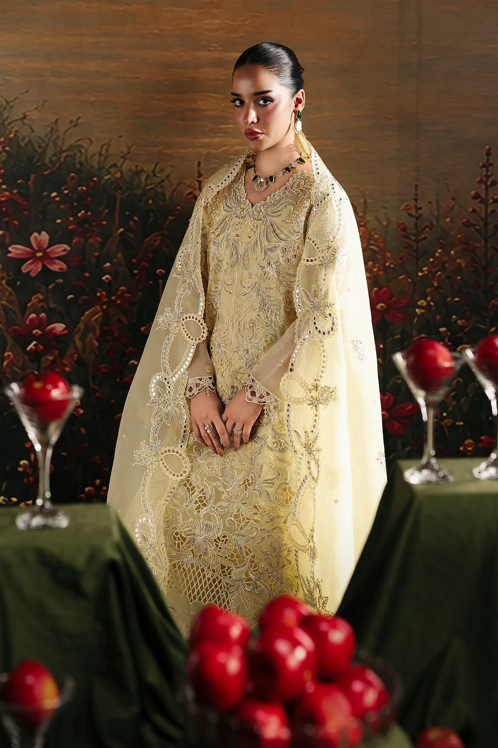 A woman models a traditional Indian outfit consisting of a cream-colored embroidered kurta and a matching sheer dupatta, set against a dark, decorative background with red apples on tables.