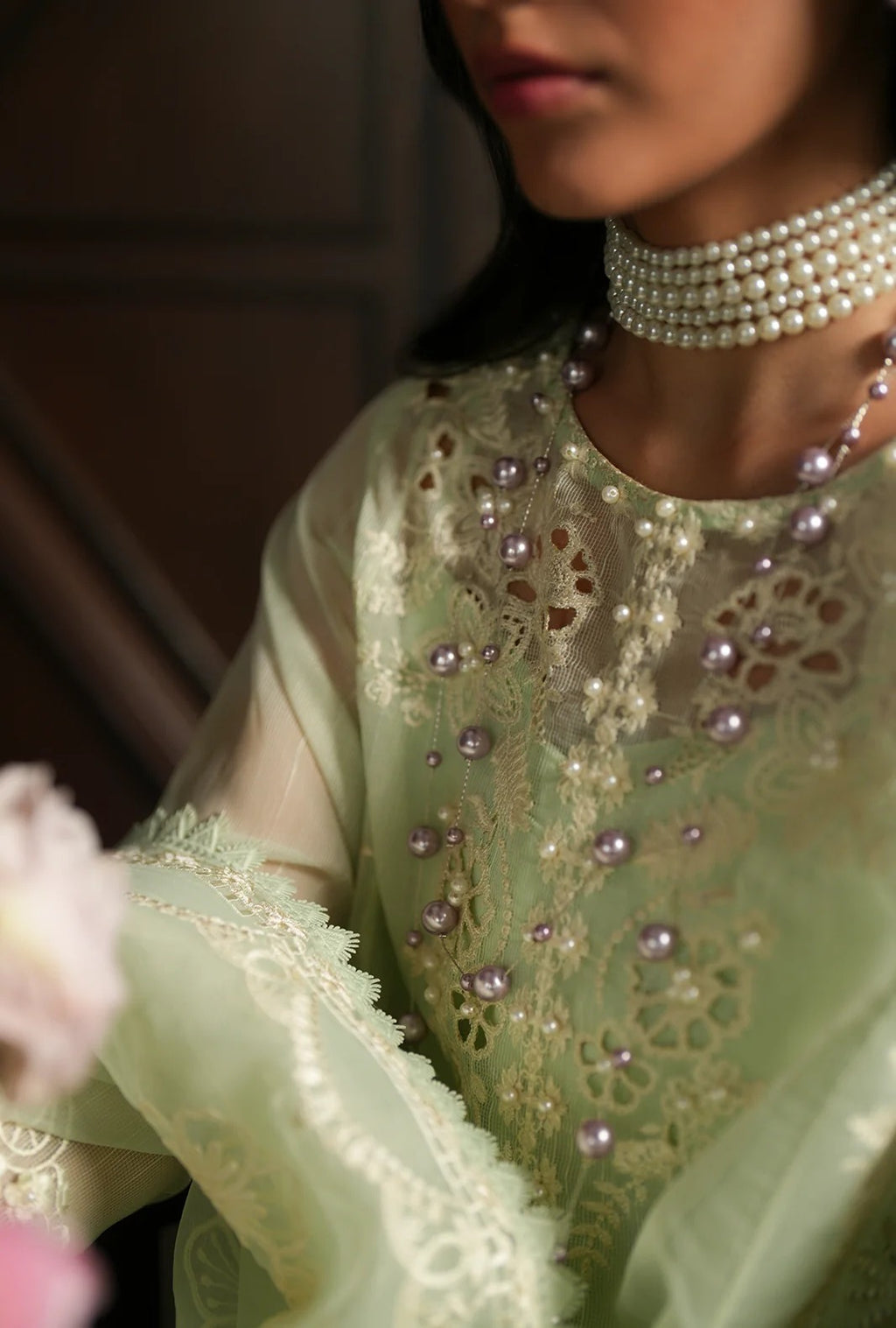 A close-up of a model wearing a light sage green embroidered traditional outfit with pearl embellishments and a matching multi-strand pearl choker necklace.