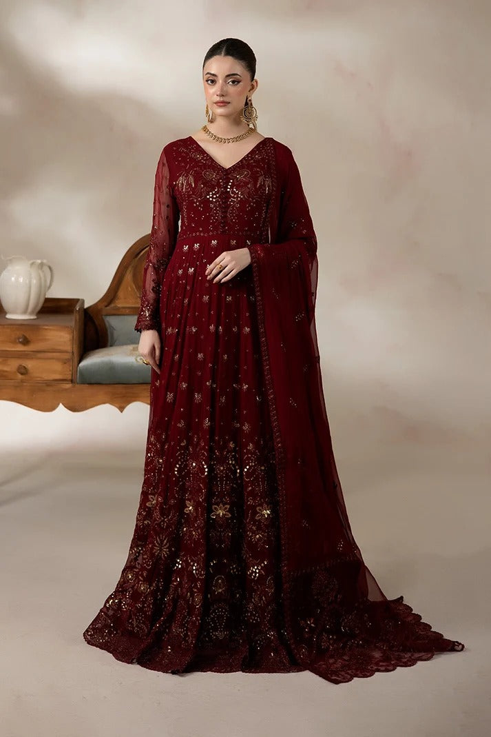 A woman models a floor-length maroon red ethnic gown with gold embroidery, sheer sleeves, and a matching dupatta, set against a warm beige studio backdrop with a wooden chair and side table.