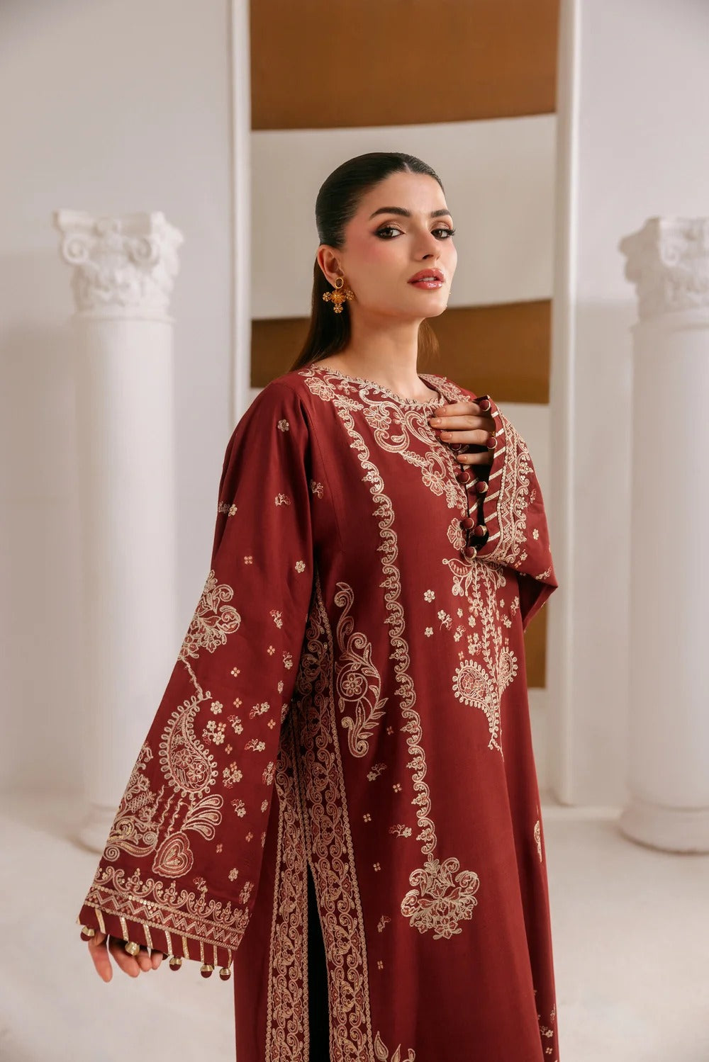 A woman modeling a long-sleeved maroon traditional tunic with intricate beige embroidery, standing in a bright, modern interior with white columns.