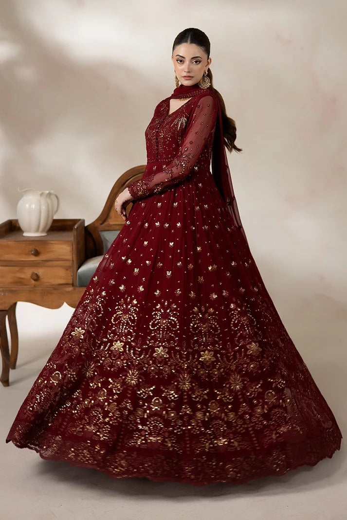A woman models a deep maroon red anarkali gown with intricate gold embroidery, set against a warm beige studio background with a wooden side table.
