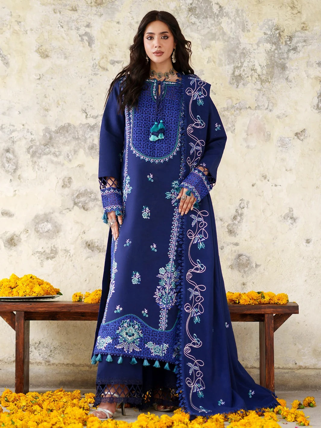 A woman models a deep navy blue traditional embroidered kurta set with matching trousers, standing in front of a rustic textured wall with yellow marigold flowers on a bench and scattered on the floor.