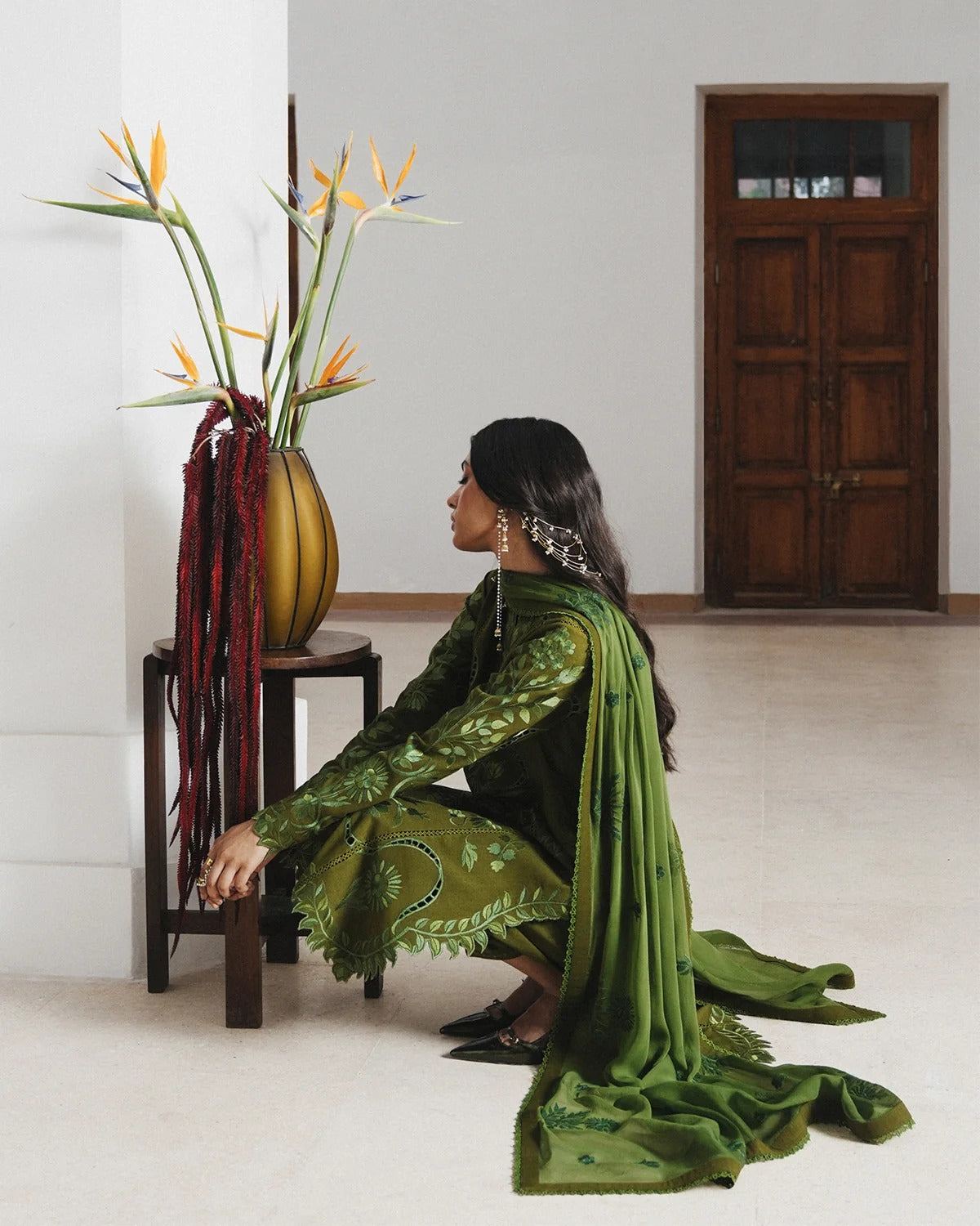 A woman in a green embroidered traditional outfit crouches beside a small wooden table holding a tall yellow vase with orange flowers, in a minimalist indoor room with a wooden door in the background.