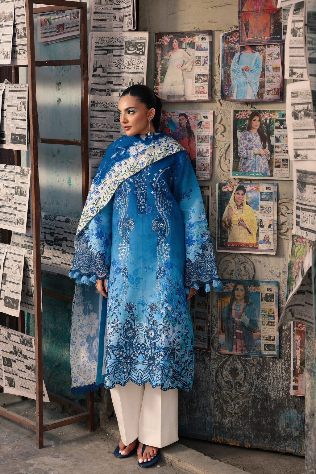A woman models a traditional blue embroidered kurta set with white pants and a matching dupatta, standing in front of a wall covered with vintage posters and newspaper clippings.