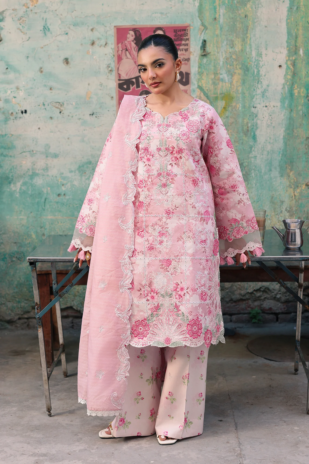 A woman models a light pink floral printed salwar kameez suit with a matching dupatta, standing in front of a rustic, weathered green wall.
