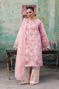 A woman models a light pink floral printed salwar kameez suit with a matching dupatta, standing in front of a rustic, weathered green wall.