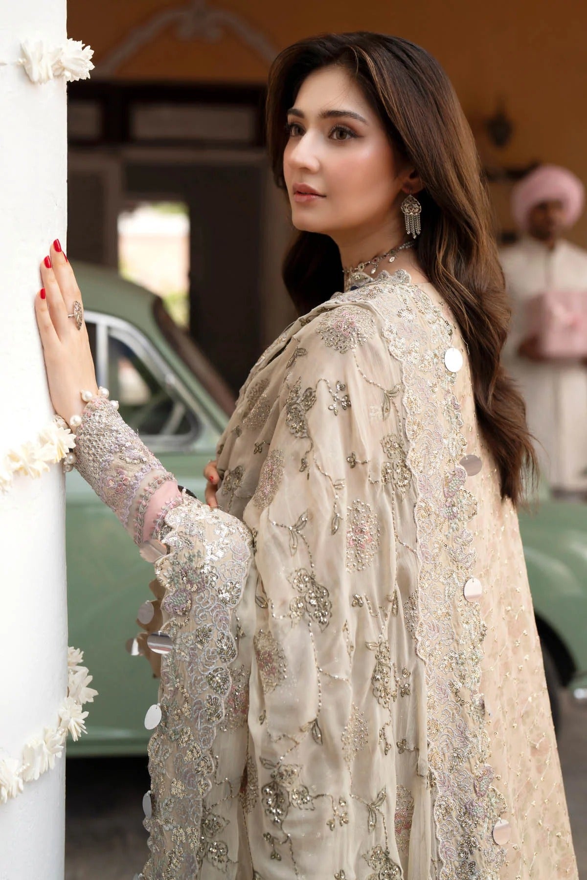 A woman wearing an ornate beige embroidered traditional outfit with a matching sheer shawl draped over her shoulder, posed beside a white pillar in an outdoor setting with a vintage green car in the background.