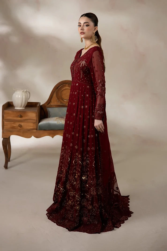 A woman models a floor-length maroon red anarkali gown with intricate gold embroidery and sheer sleeves, posed in front of a neutral studio backdrop with a wooden bench prop.
