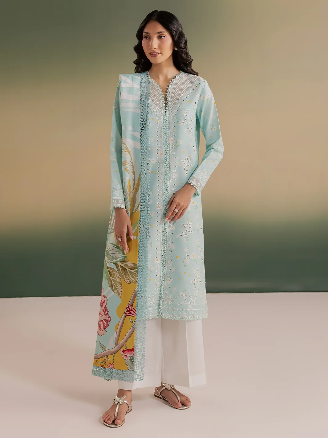 A woman modeling a light blue-green embroidered kurta with a floral print panel, paired with white pants and sandals, set against a gradient studio background.