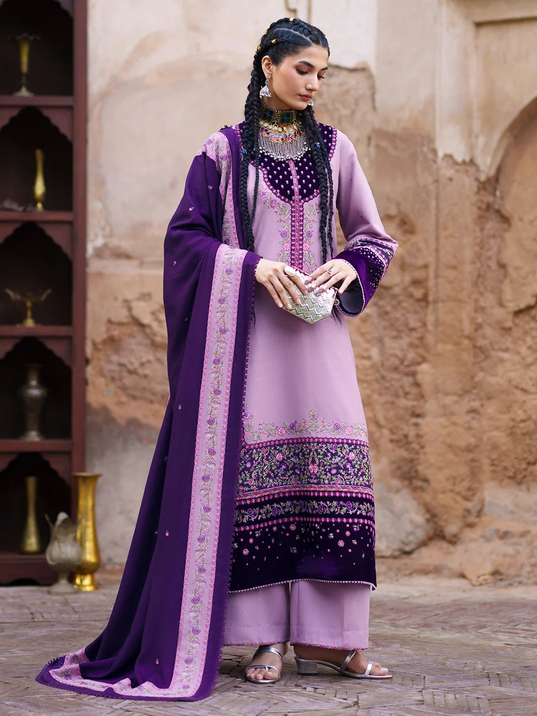 A woman models a traditional South Asian salwar kameez suit in lavender purple and deep purple. The outfit features a lavender kurta with a deep purple border, matching lavender trousers, and a deep purple dupatta with intricate embroidery.