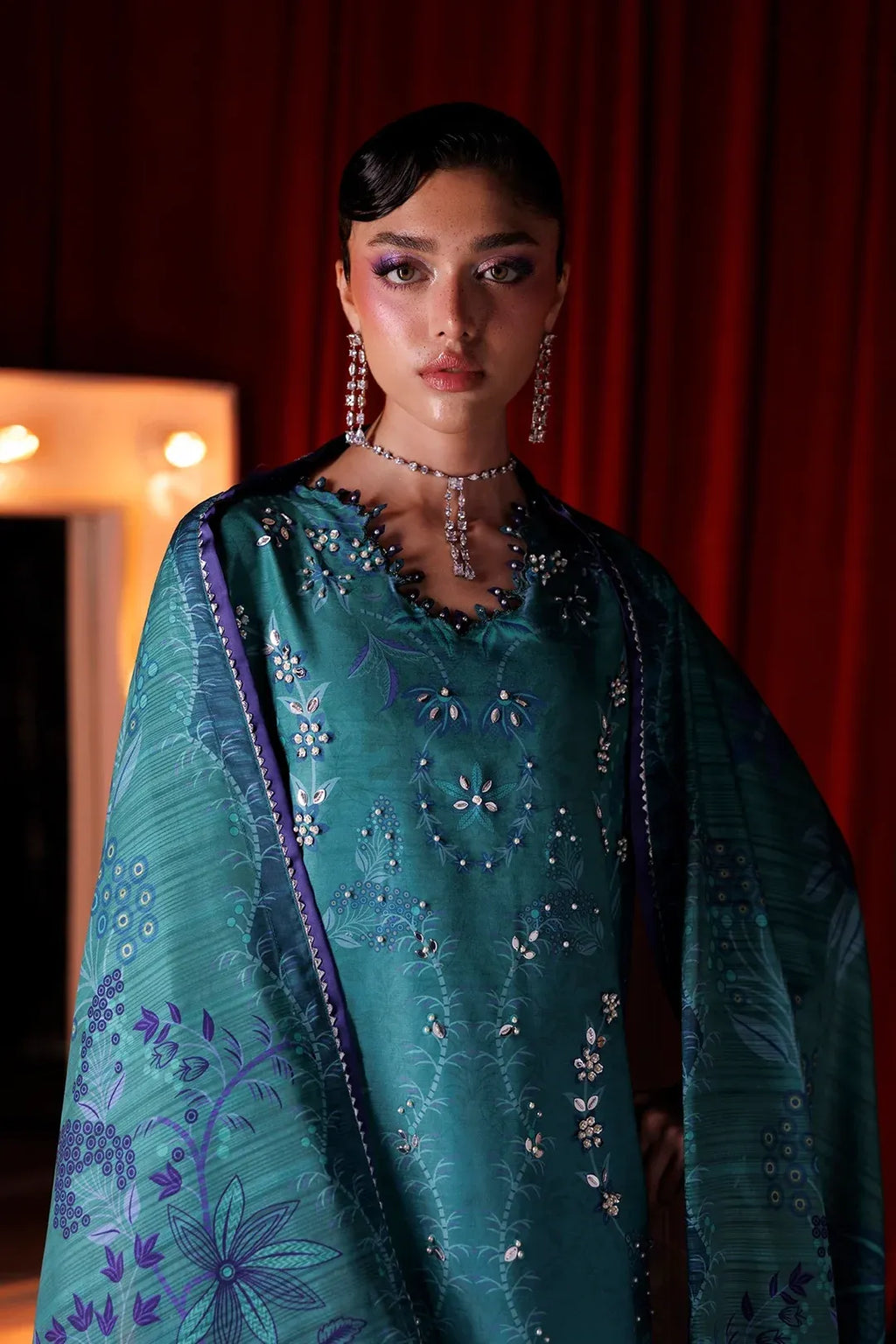A model wearing a teal green embroidered traditional outfit with a matching sheer dupatta, set against a dark red curtain background.
