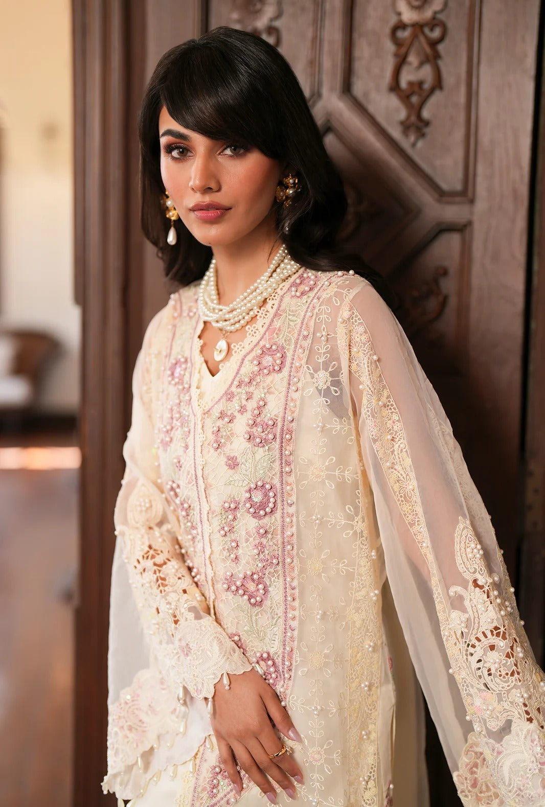 A woman wearing a cream-colored traditional embroidered outfit with pink floral embroidery and sheer sleeves, standing in front of a dark wooden door.