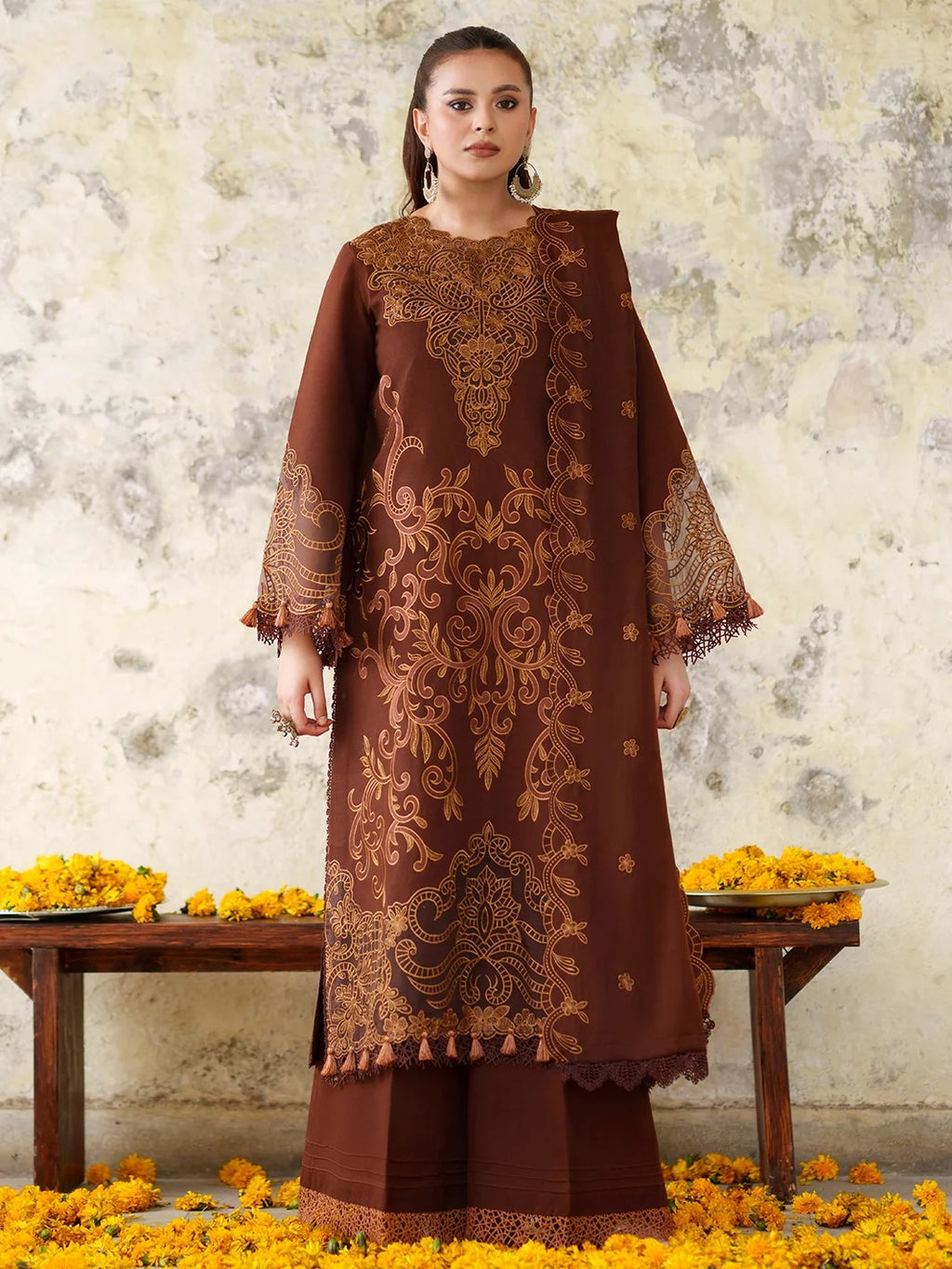 A woman models a brown traditional kurta set with intricate gold embroidery, paired with matching brown wide-leg trousers, set against a rustic textured wall with yellow flowers on a bench.