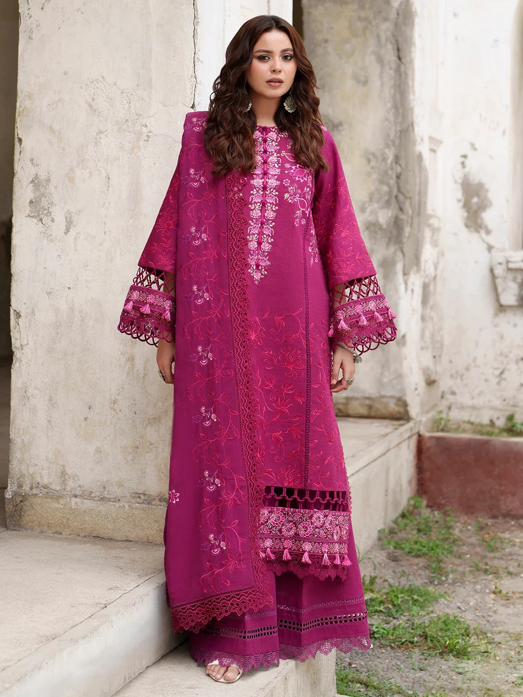 A woman modeling a magenta pink embroidered kurta set with matching trousers, standing on stone steps against a weathered concrete wall.