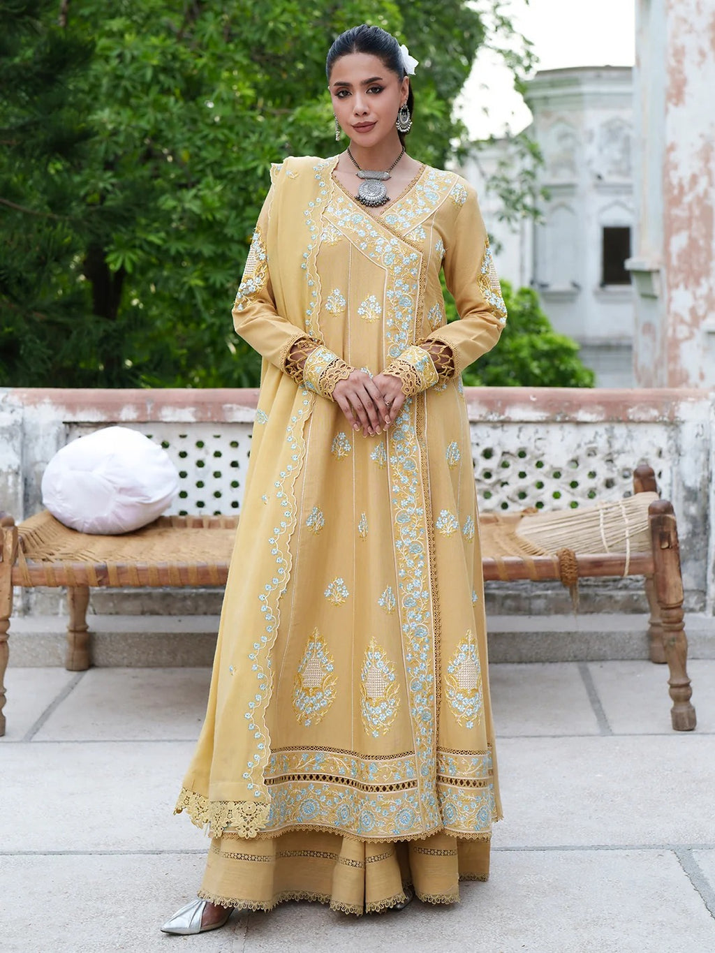 A woman stands outdoors wearing a long mustard yellow embroidered Anarkali gown with matching pants and silver flat shoes. The dress has light blue and white floral motifs, a V-neckline, and ornate lace borders.