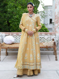 A woman stands outdoors wearing a long mustard yellow embroidered Anarkali gown with matching pants and silver flat shoes. The dress has light blue and white floral motifs, a V-neckline, and ornate lace borders.