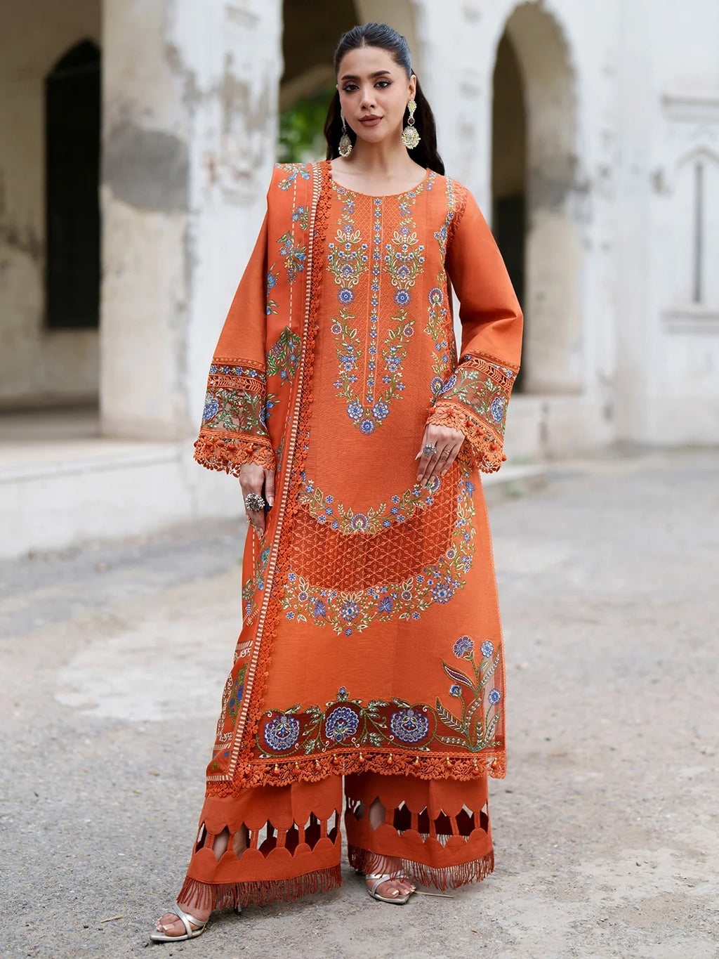 A woman stands outdoors wearing a bright orange embroidered traditional outfit with matching wide-leg pants and a draped scarf. The outfit features intricate multicolor floral and geometric embroidery in blue, green, and gold tones, with lace trim and fringe details.