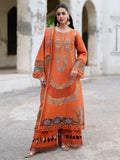 A woman stands outdoors wearing a bright orange embroidered traditional outfit with matching wide-leg pants and a draped scarf. The outfit features intricate multicolor floral and geometric embroidery in blue, green, and gold tones, with lace trim and fringe details.