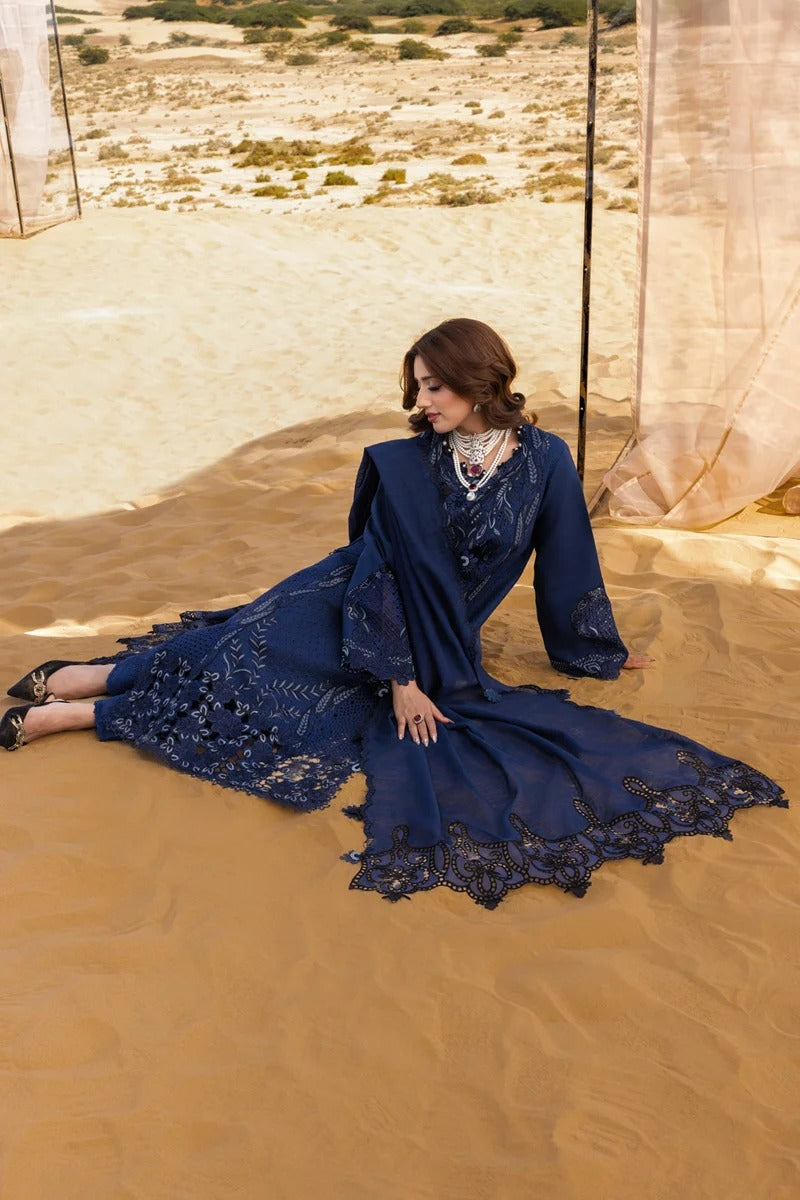 A woman wearing a navy blue embroidered traditional dress sits on a sandy desert dune under sheer beige fabric drapes.