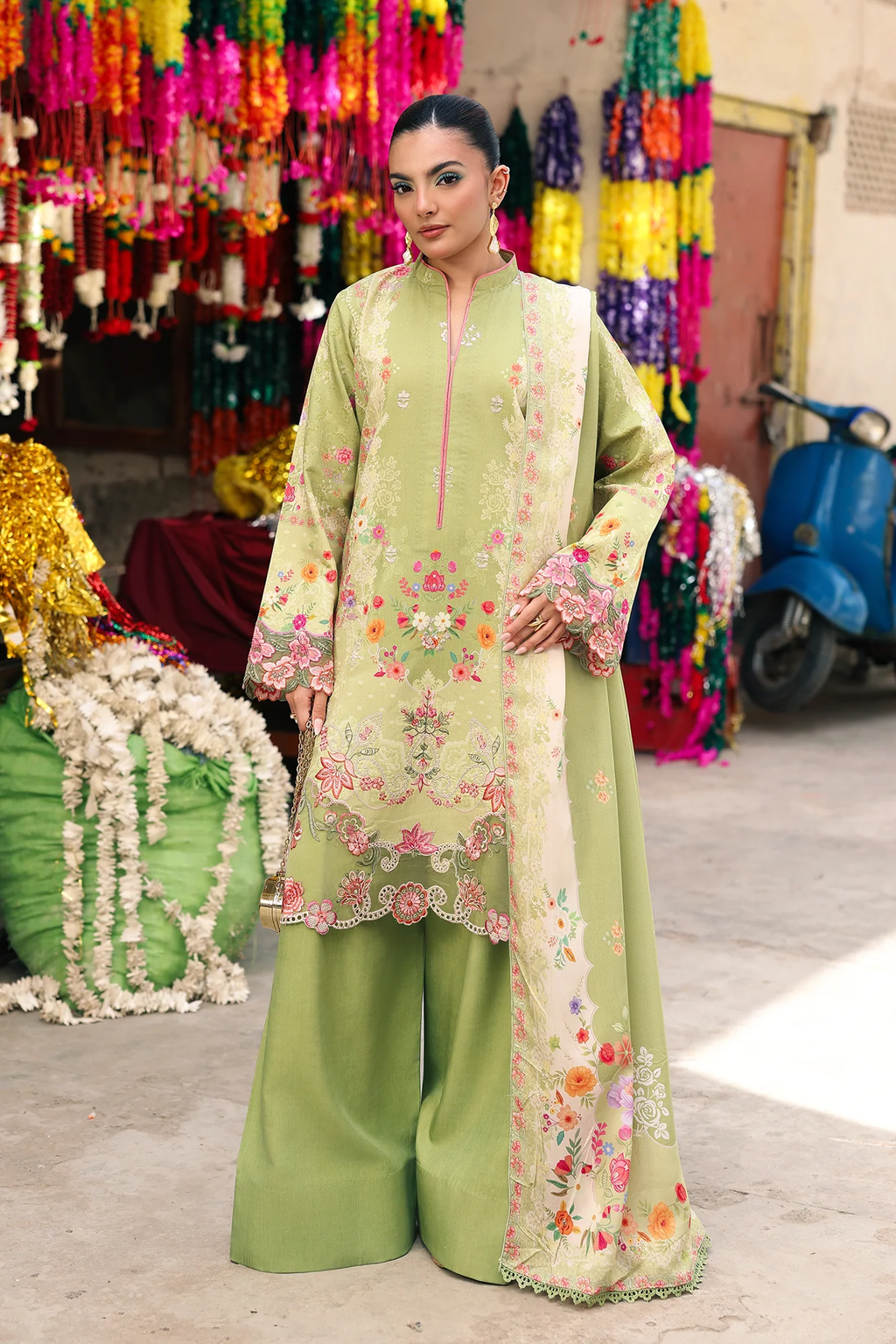 A woman models a light olive green three-piece ethnic suit with pink and multicolor floral embroidery, standing in a market setting with colorful hanging decorations and a blue scooter in the background.