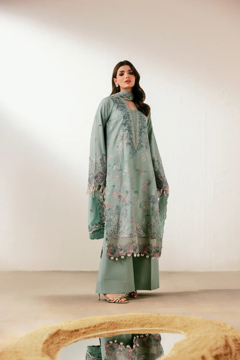 A woman models a traditional three-piece salwar kameez suit in sage green with intricate floral embroidery and beadwork on the tunic, paired with matching wide-leg trousers and heels, set against a minimalist beige studio background.