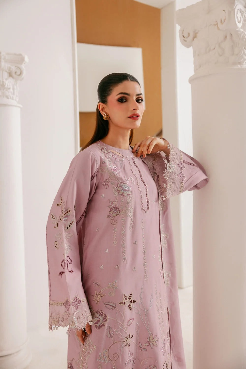 A woman models a light pink embroidered traditional outfit with long sleeves and lace-trimmed cuffs, standing between white columns in an elegant indoor setting.