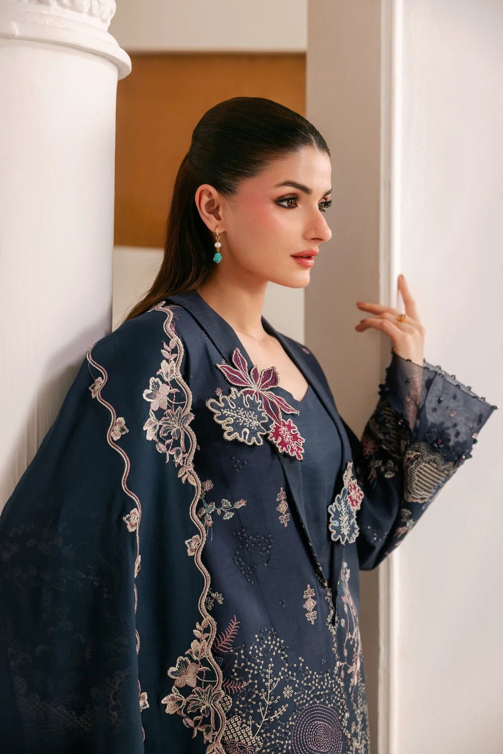 A woman models a dark navy blue embroidered traditional outfit with intricate floral patterns and sheer sleeves, posing next to a white architectural column.
