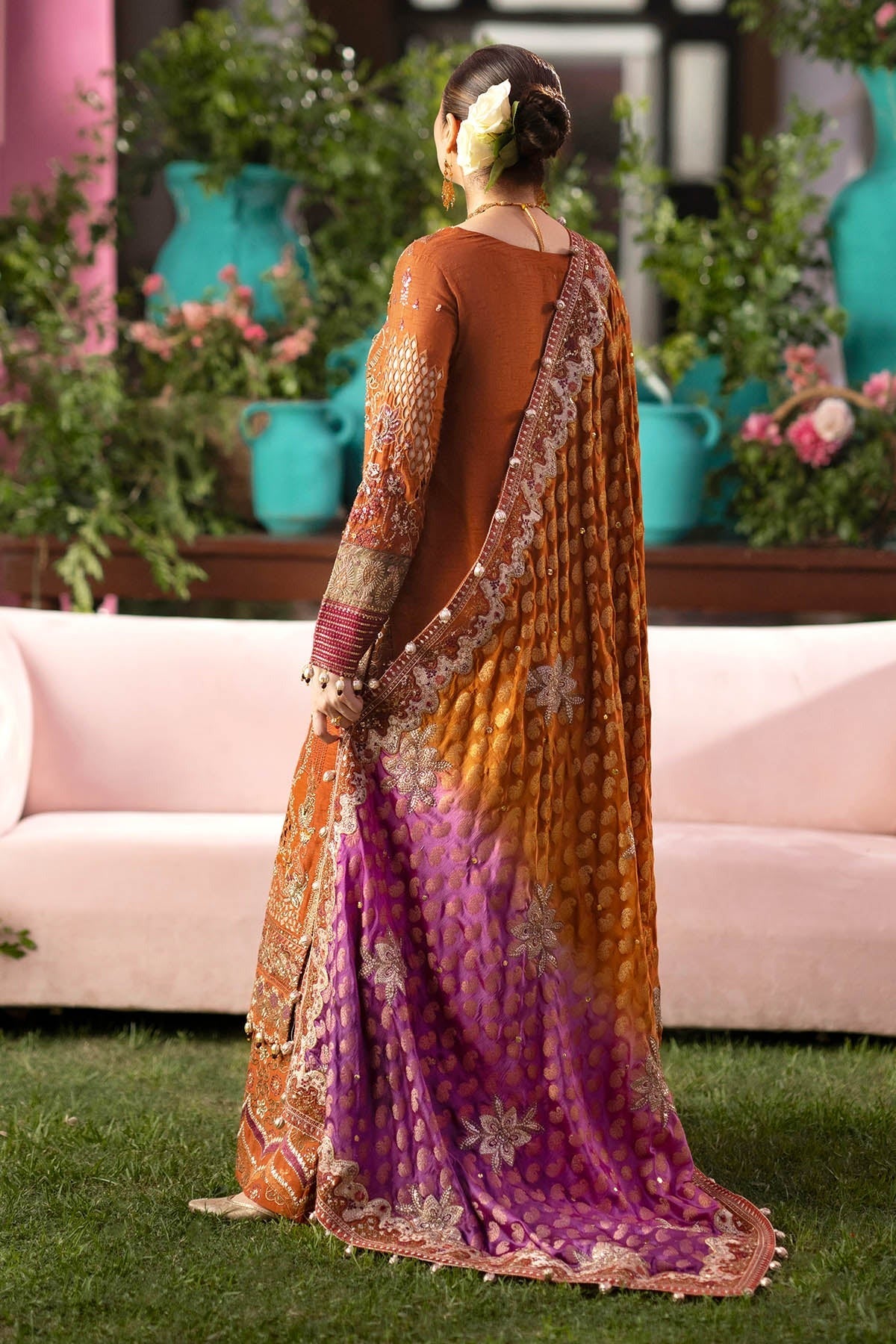A woman stands outdoors on grass in front of a pink sofa, wearing a traditional draped outfit with a warm orange-brown top and a long patterned drape that fades into purple at the bottom, decorated with metallic floral motifs.