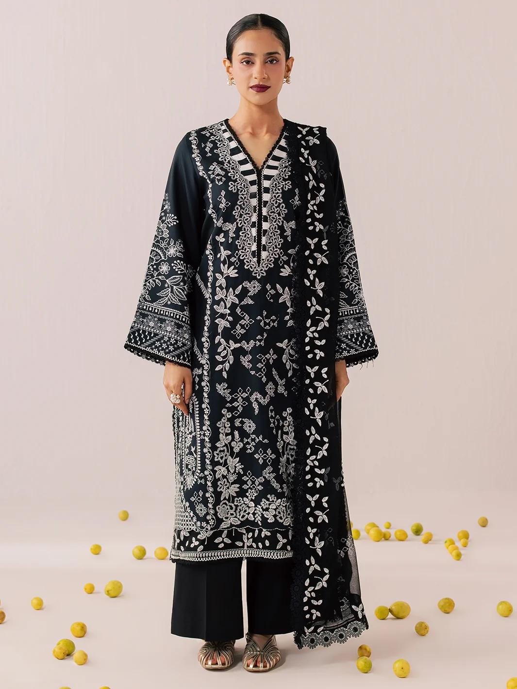 A woman modeling a black embroidered kurta with white floral patterns, paired with black pants, standing against a light beige background with scattered yellow lemons on the floor.