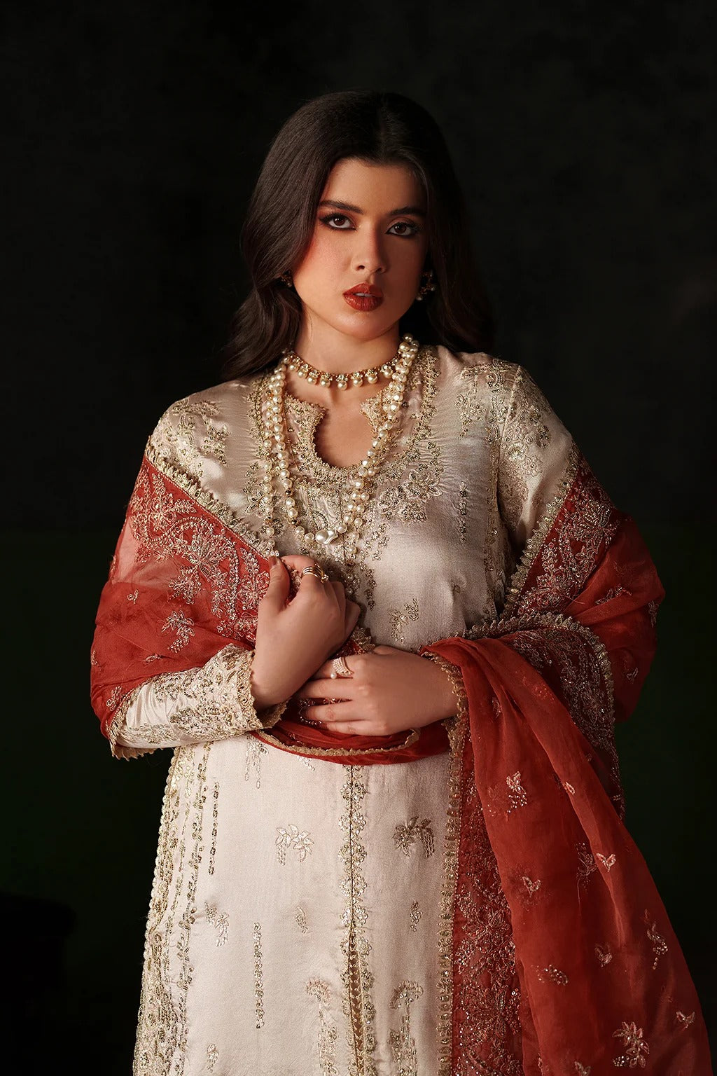 A woman modeling a traditional South Asian outfit featuring a cream-colored embroidered kurta, a rust-red dupatta with gold floral patterns, and a matching pearl necklace, set against a dark studio background.