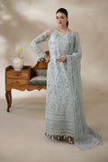 A woman models a light blue-gray traditional embroidered dress with sheer sleeves and a matching dupatta, standing in a studio with a vintage wooden side table and chair in the background.