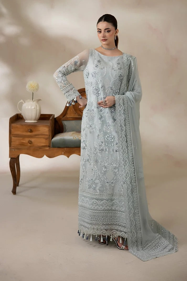 A woman models a light blue-gray traditional embroidered dress with sheer sleeves and a matching dupatta, standing in a studio with a vintage wooden side table and chair in the background.