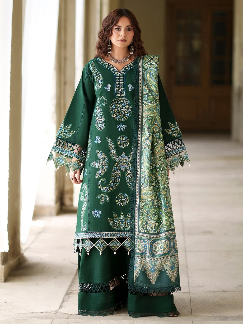 A woman modeling a dark green traditional South Asian outfit with intricate gold, blue, and white embroidery, standing in a softly lit corridor.
