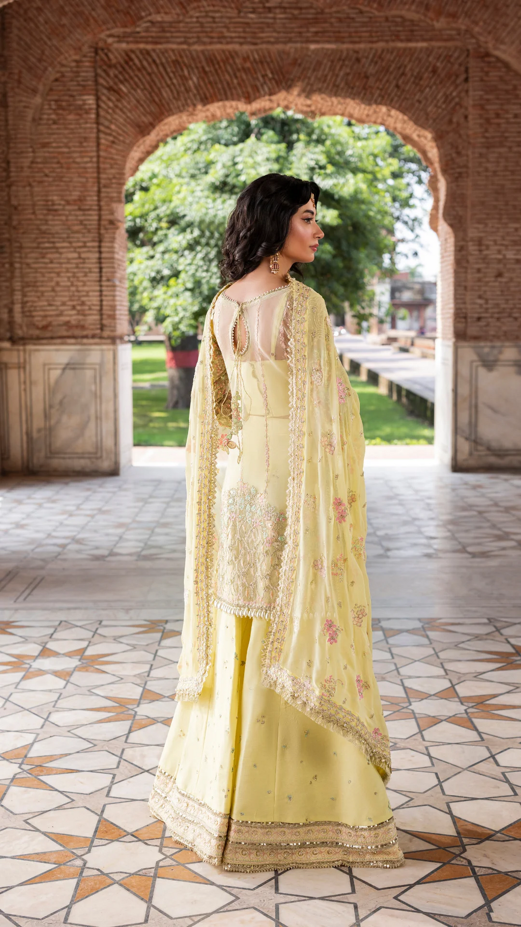 Woman in a light yellow traditional outfit standing in front of an archway with greenery outside.
