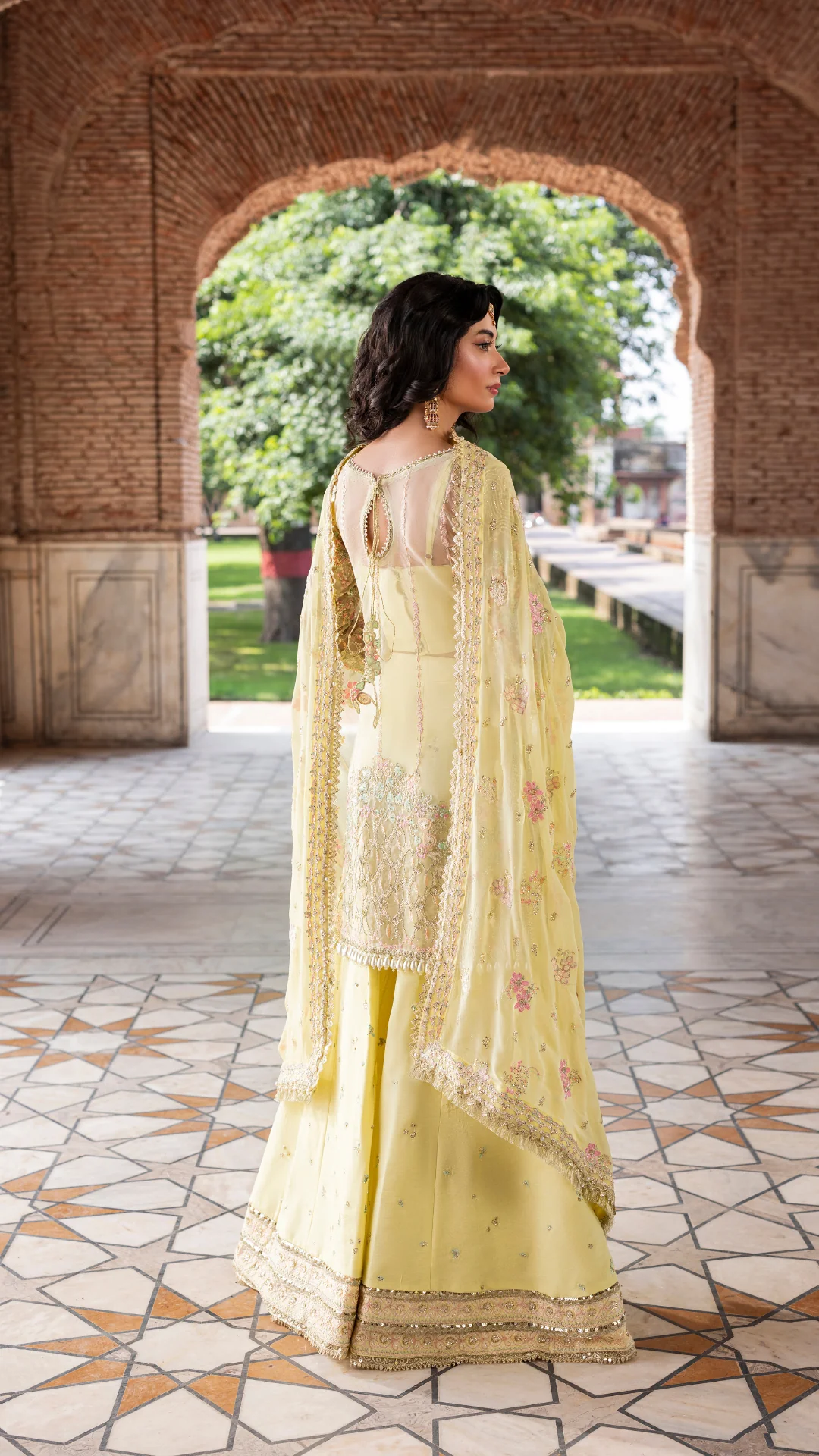 Woman in a light yellow traditional outfit standing in front of an archway with greenery outside.