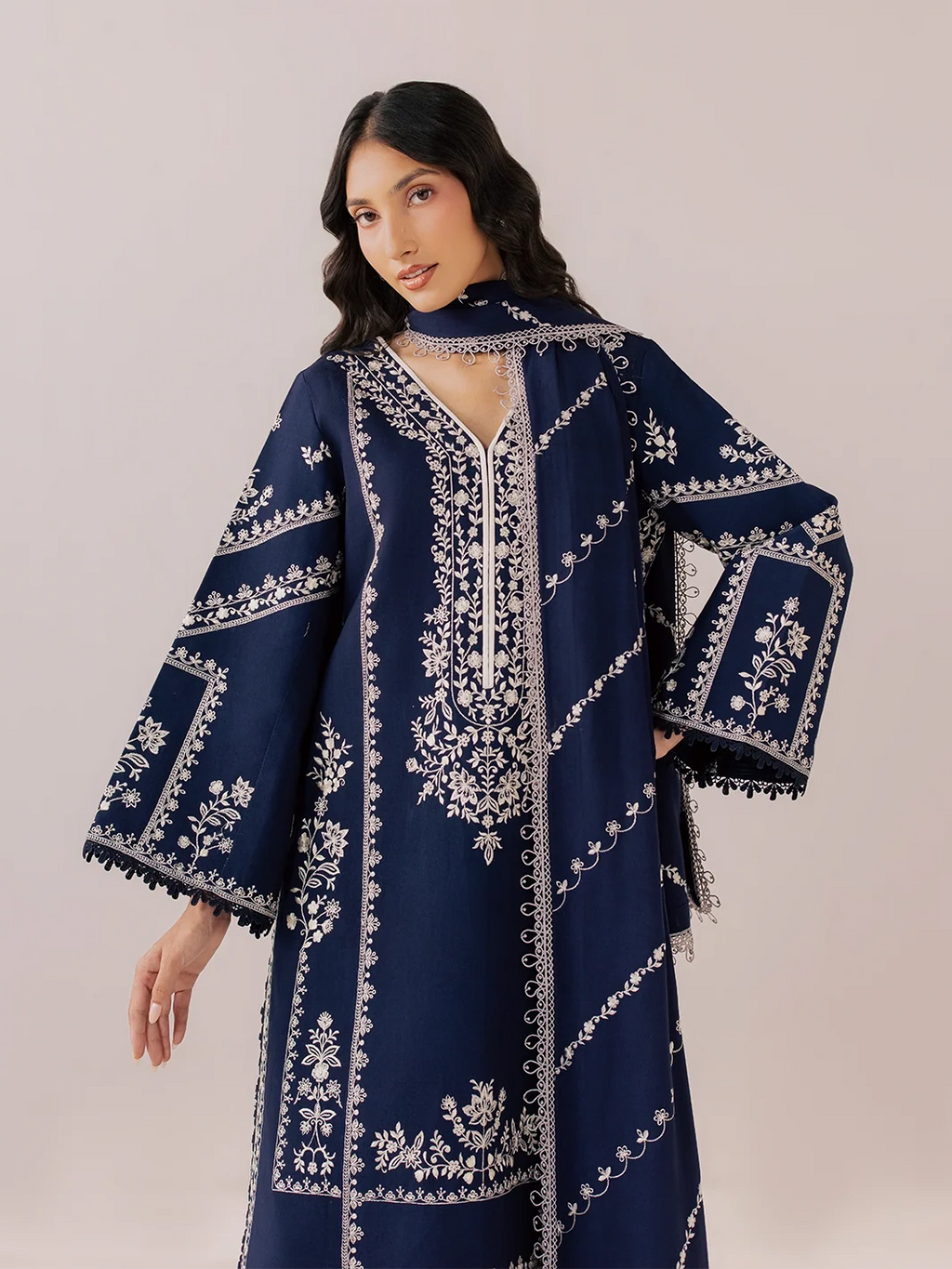 A woman modeling a navy blue kaftan dress with intricate white embroidery, featuring wide sleeves, a high collar, and a split neckline, set against a solid light beige background.