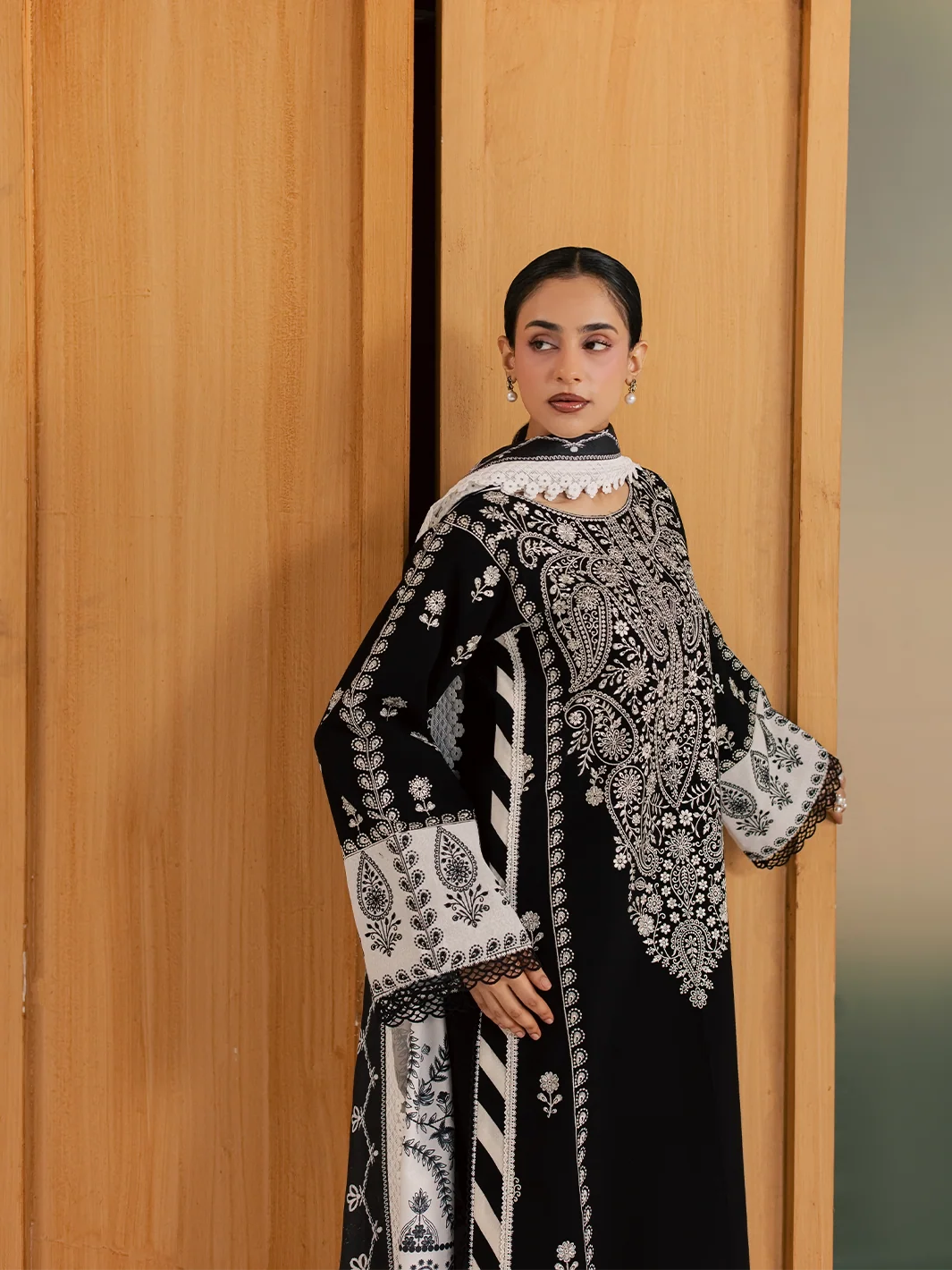 A woman models a black traditional embroidered outfit with intricate white and gray floral patterns, standing in front of a light wood-paneled wall.