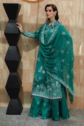 Woman in a teal embroidered outfit standing against a beige wall.