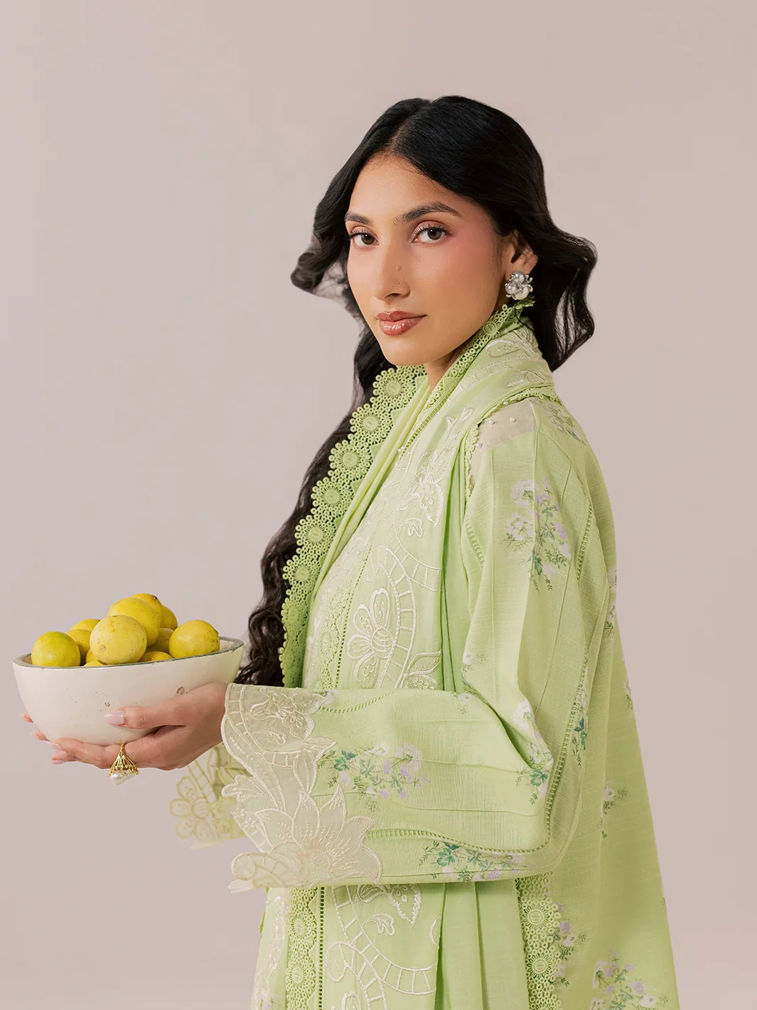 A woman wearing a light green embroidered traditional outfit holds a white bowl filled with lemons against a solid pale pink background.