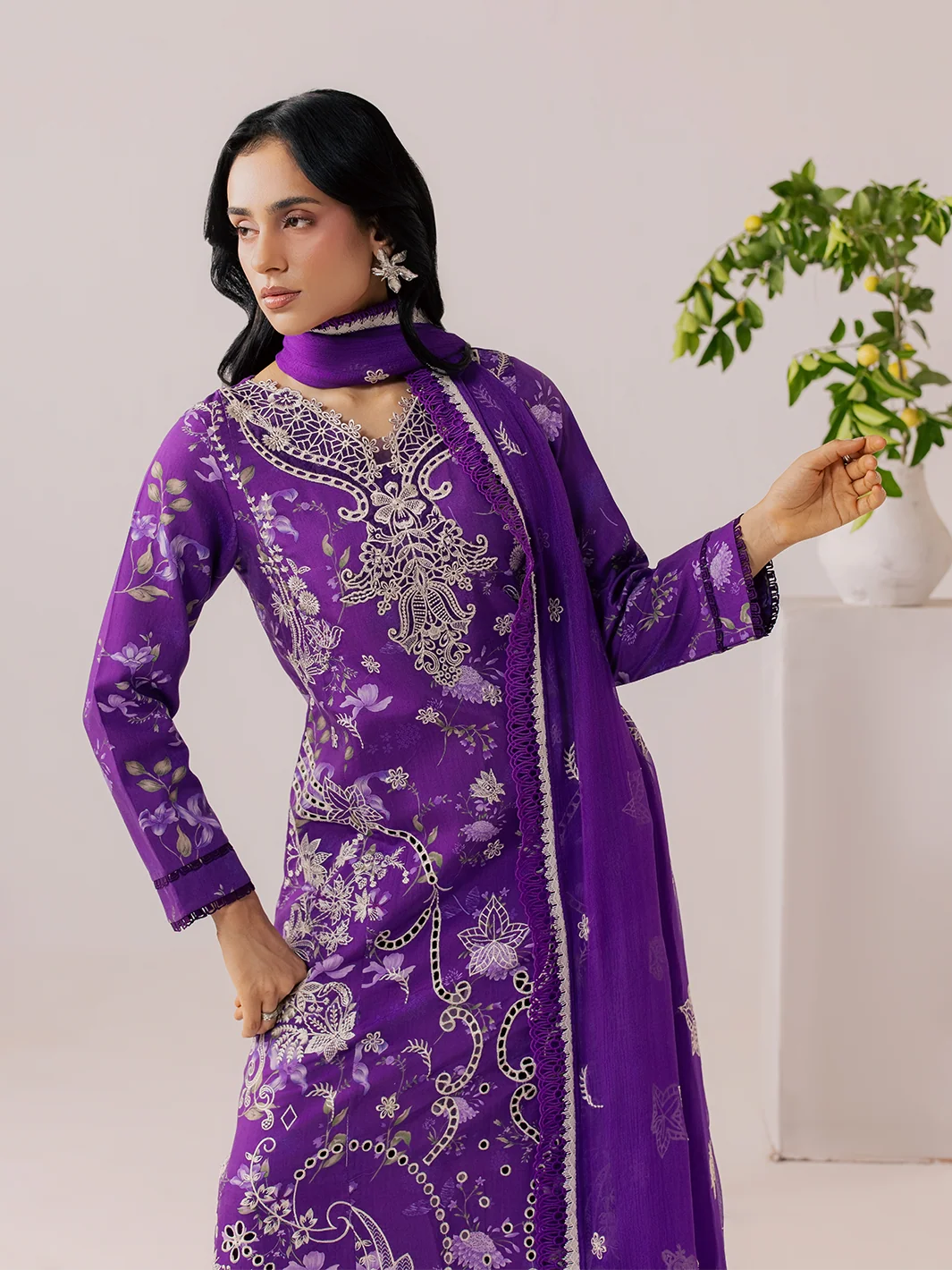 A woman models a purple traditional outfit with intricate silver and white floral embroidery, featuring a long-sleeved kurta, matching high-neck choker, and a coordinating dupatta, set against a light beige background with a decorative plant.