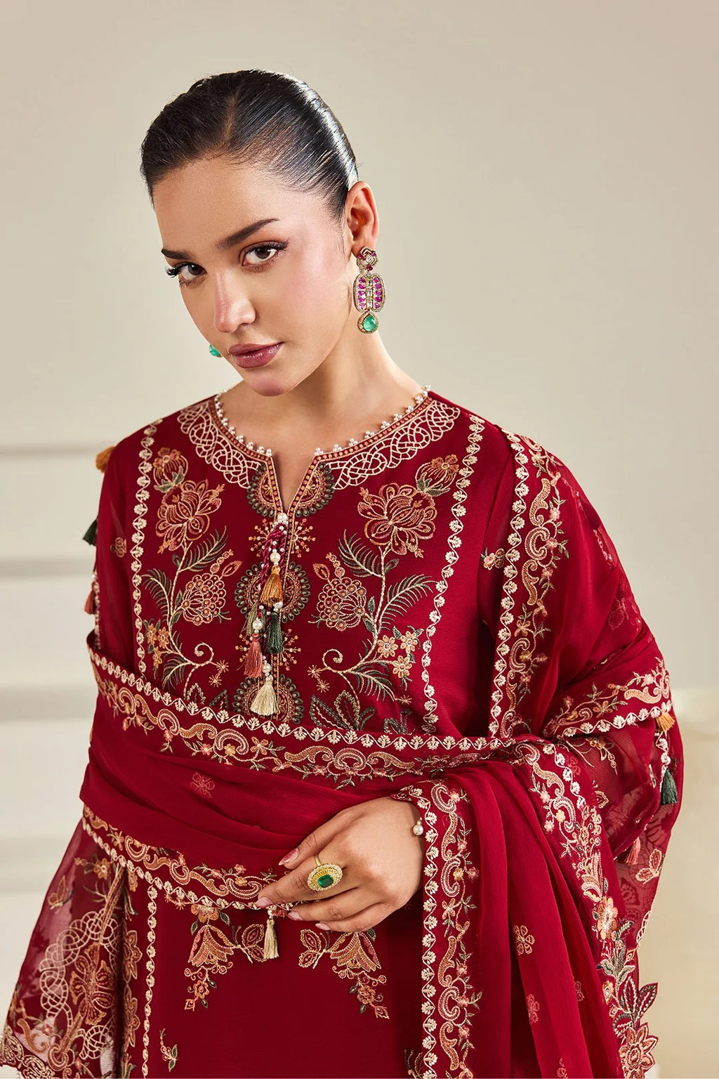 A woman wearing a deep red embroidered traditional outfit with intricate gold and beige floral detailing, paired with a matching draped dupatta, against a neutral beige background.