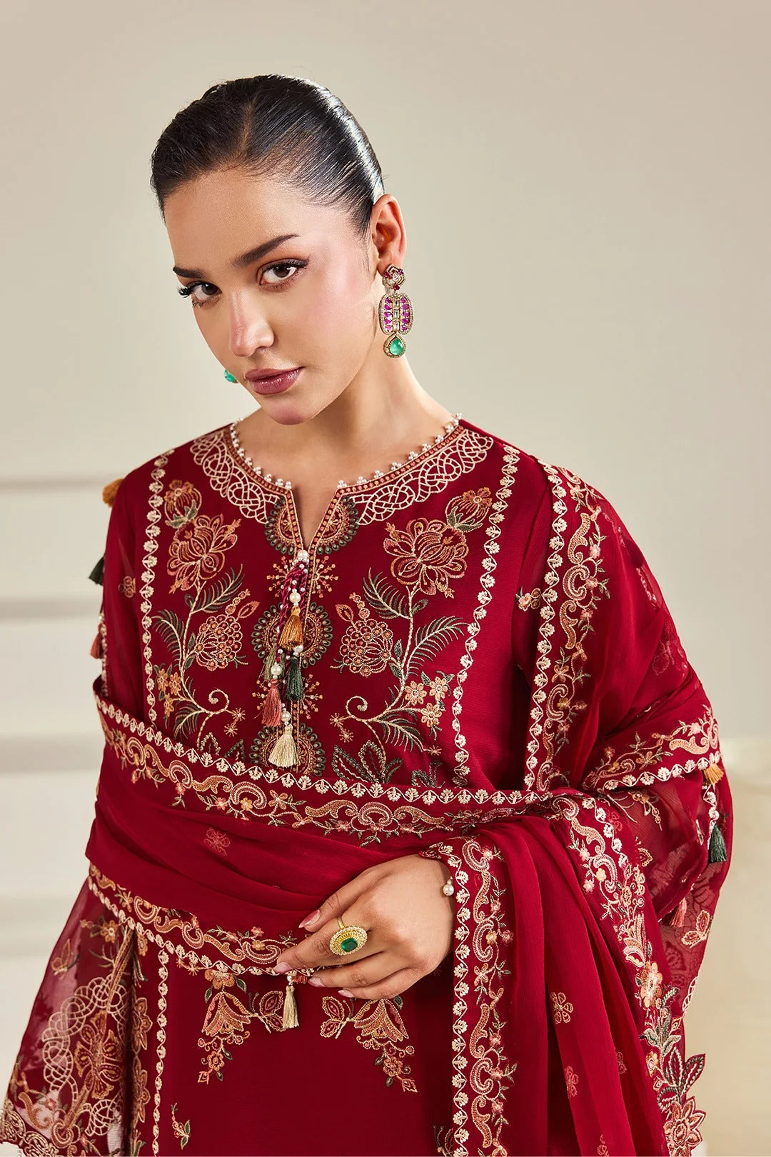 A woman wearing a deep red embroidered traditional outfit with intricate gold and beige floral detailing, paired with a matching draped dupatta, against a neutral beige background.