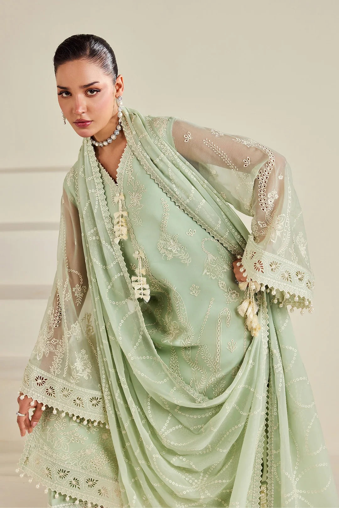 A woman models a light sage green traditional outfit with matching sheer dupatta, featuring intricate gold embroidery, white lace borders, and tassel details against a neutral beige background.