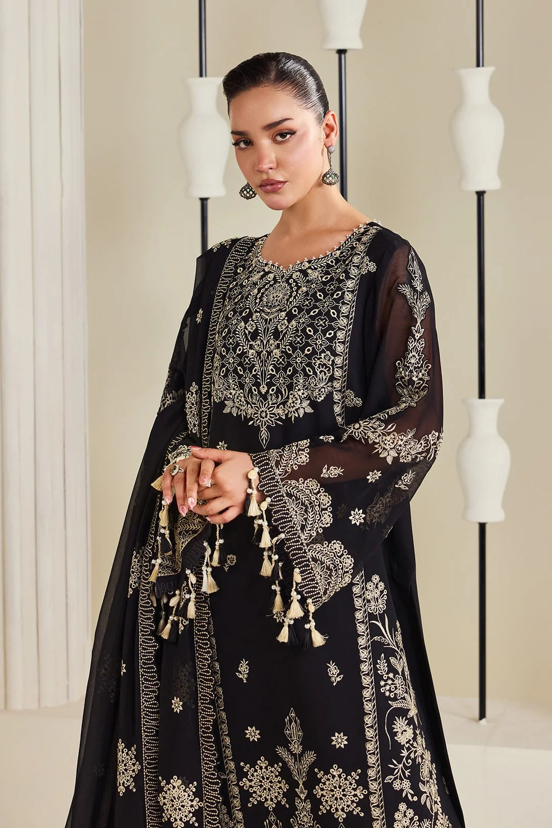 A woman models a black embroidered traditional outfit with beige floral patterns and tassel details, set against a modern indoor backdrop with decorative white vases.