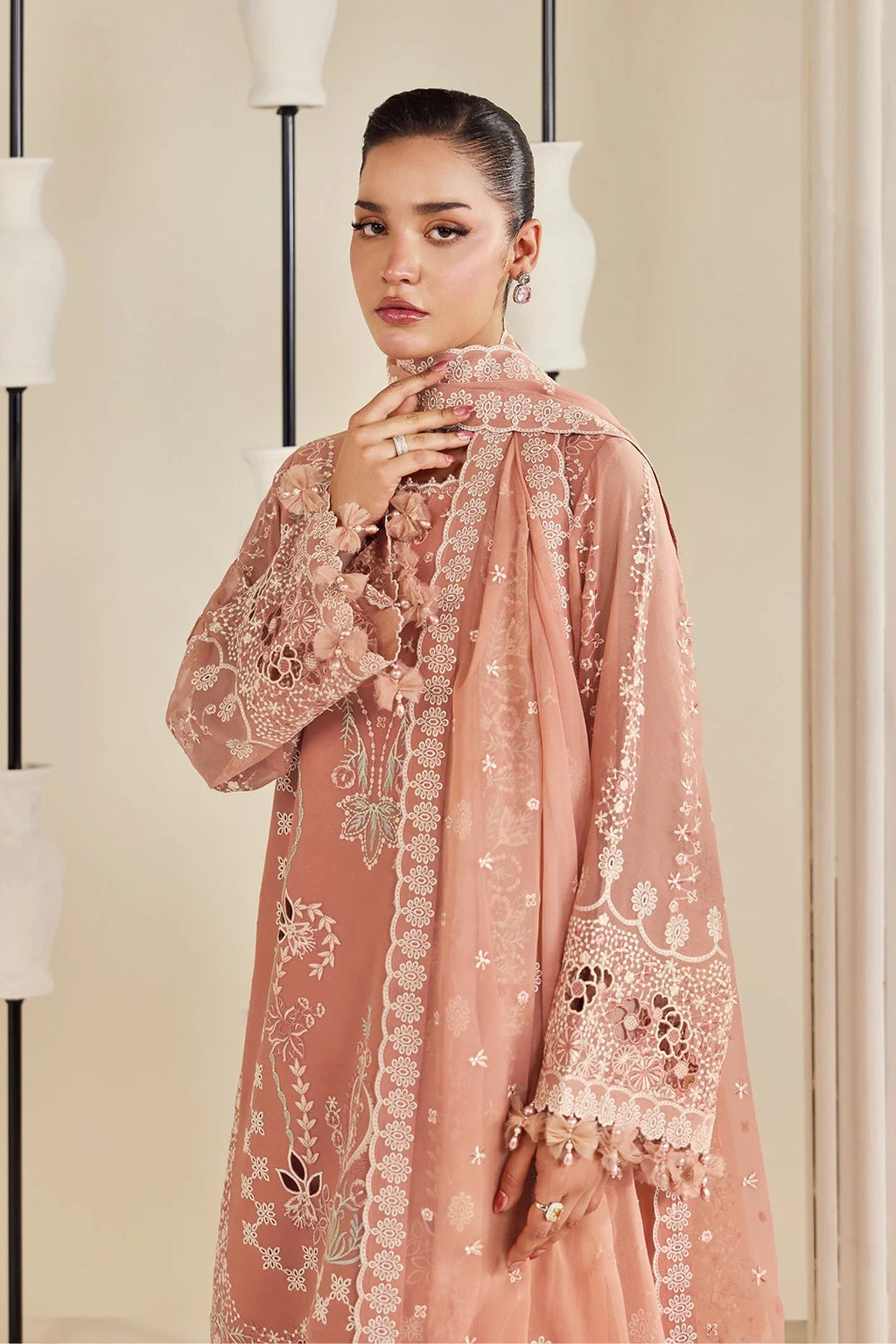 A woman models a traditional three-piece salwar kameez suit in dusty rose pink, featuring intricate white and gold floral embroidery on the kurta and dupatta, set against a warm beige background with modern minimalist lamp posts.