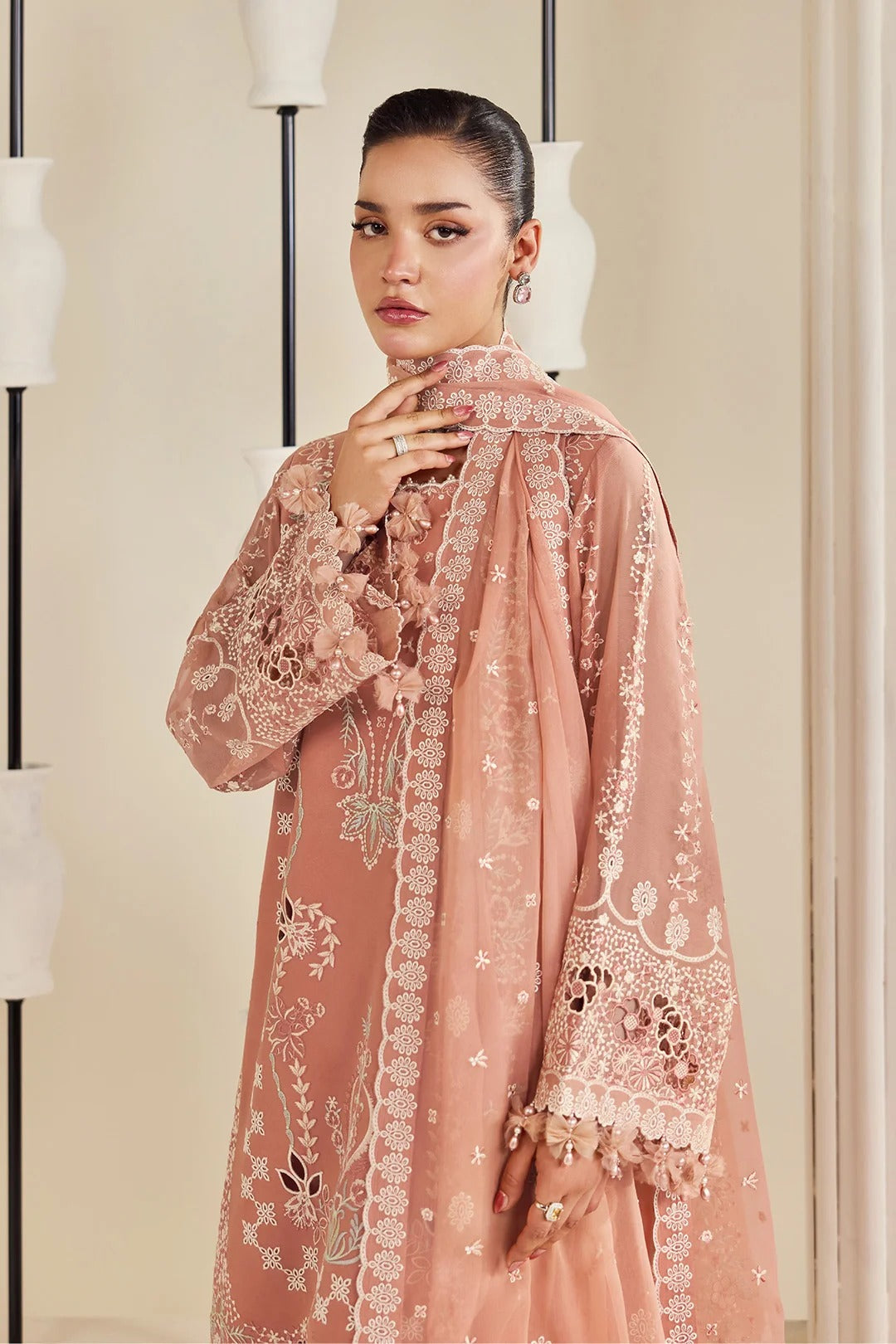 A woman models a traditional three-piece salwar kameez suit in dusty rose pink, featuring intricate white and gold floral embroidery on the kurta and dupatta, set against a warm beige background with modern minimalist lamp posts.