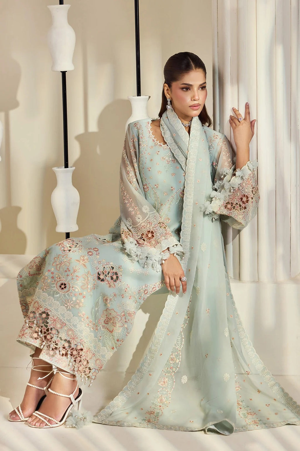 A woman models a light blue-green embroidered traditional outfit with a matching scarf and sheer sleeves, posed in a styled interior with decorative white vases and draped curtains.
