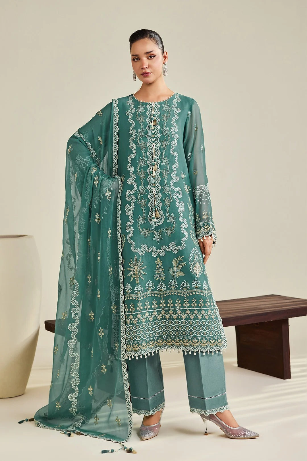A woman models a traditional three-piece teal green ethnic suit with intricate gold and silver embroidery, featuring a long kurta, matching trousers, and a sheer dupatta, set against a minimalist beige background with a wooden bench.