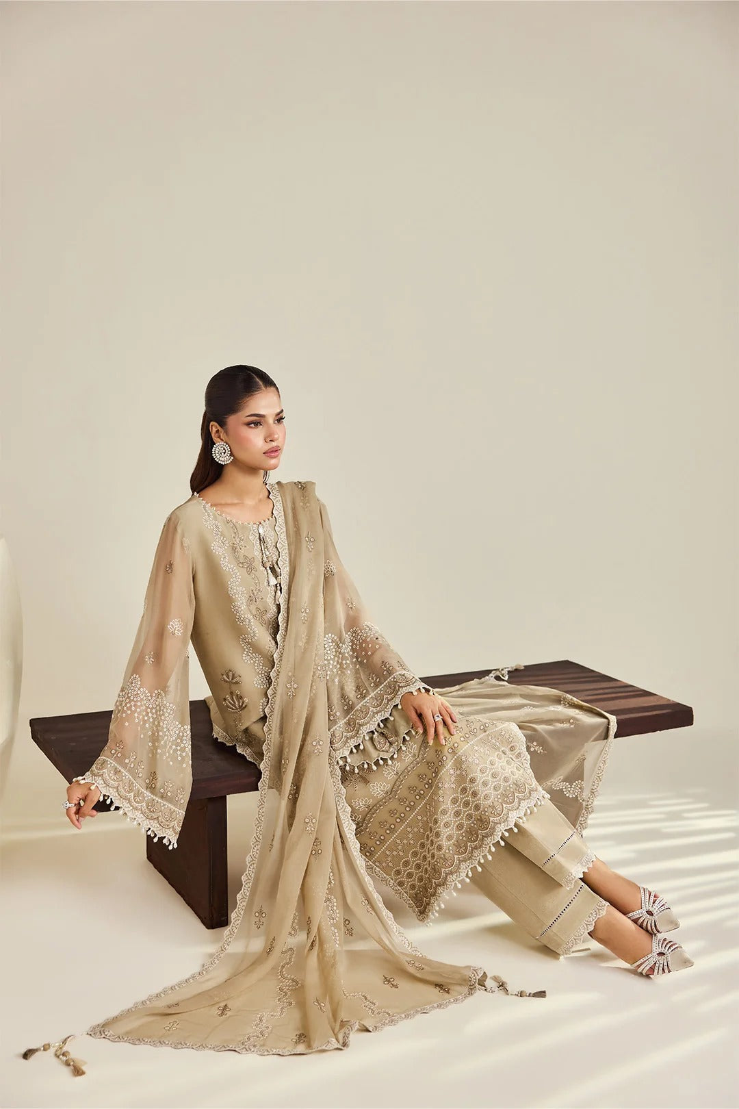 A woman models a beige three-piece traditional outfit with matching dupatta, seated on a dark wooden bench against a warm off-white studio background.
