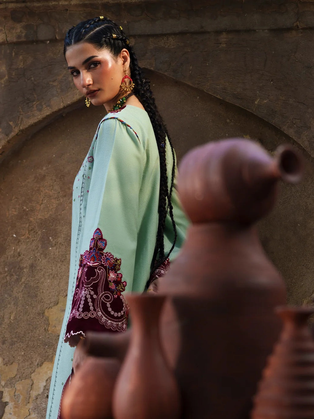 A woman wearing a mint green traditional outfit with dark purple embroidered sleeve details, standing in front of a rustic stone archway with clay pots in the foreground.